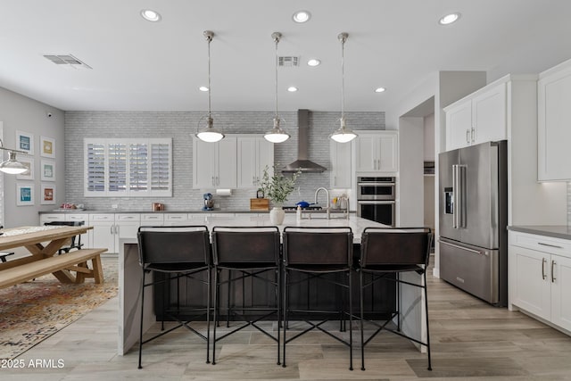 kitchen with pendant lighting, stainless steel appliances, an island with sink, and wall chimney range hood