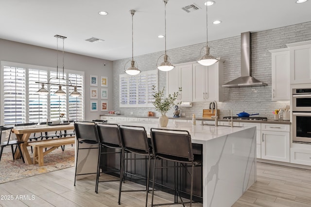 kitchen featuring white cabinetry, hanging light fixtures, stainless steel appliances, an island with sink, and wall chimney exhaust hood