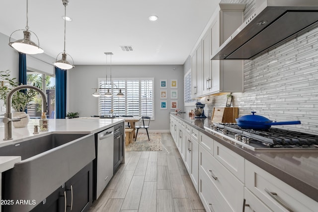 kitchen with wall chimney range hood, sink, appliances with stainless steel finishes, white cabinetry, and hanging light fixtures