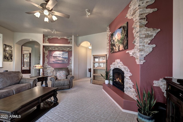 carpeted living room featuring arched walkways, a stone fireplace, and ceiling fan