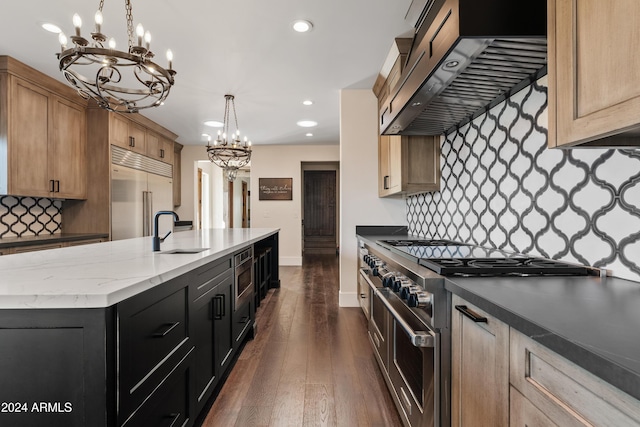 kitchen with built in appliances, dark cabinets, a sink, wall chimney range hood, and pendant lighting