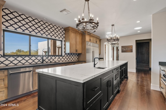 kitchen with a center island with sink, dark cabinets, decorative light fixtures, built in appliances, and a sink