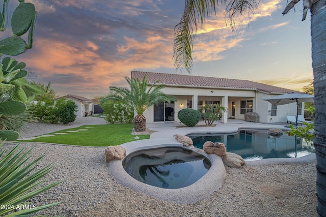 back house at dusk featuring a lawn, a swimming pool with hot tub, and a patio