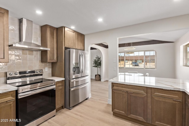 kitchen with light wood-style flooring, tasteful backsplash, arched walkways, appliances with stainless steel finishes, and wall chimney exhaust hood