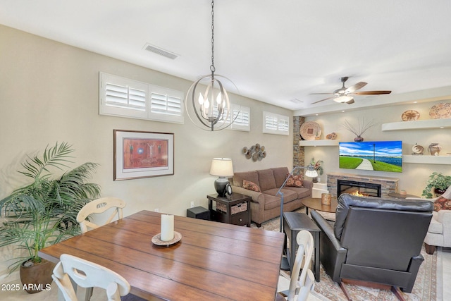 dining room with a stone fireplace and ceiling fan with notable chandelier