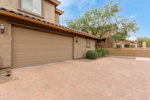 view of property exterior featuring stucco siding, decorative driveway, fence, a tile roof, and an attached garage