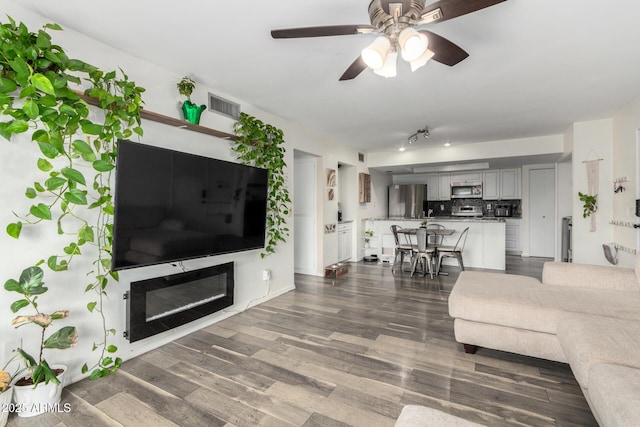 living area featuring ceiling fan, dark wood-type flooring, a glass covered fireplace, and visible vents