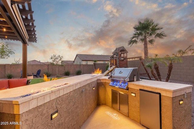 patio terrace at dusk with area for grilling, a fenced backyard, and exterior kitchen