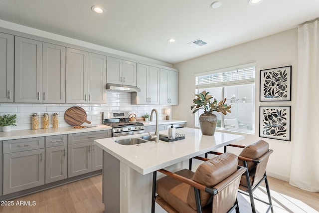 kitchen with gray cabinets, a sink, under cabinet range hood, stainless steel range with gas cooktop, and light wood-type flooring