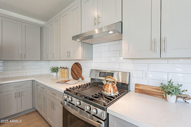 kitchen with stainless steel range with gas stovetop, under cabinet range hood, gray cabinetry, backsplash, and light stone counters