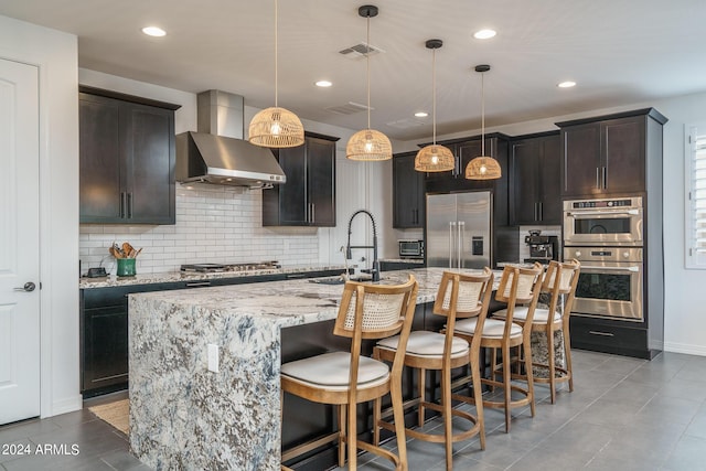 kitchen featuring a kitchen bar, wall chimney exhaust hood, decorative light fixtures, an island with sink, and stainless steel appliances