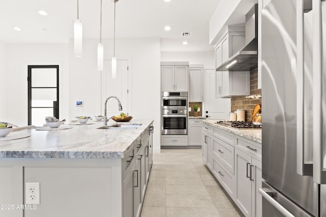 kitchen with sink, hanging light fixtures, appliances with stainless steel finishes, an island with sink, and wall chimney range hood