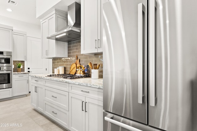 kitchen with wall chimney range hood, appliances with stainless steel finishes, backsplash, light stone counters, and white cabinets