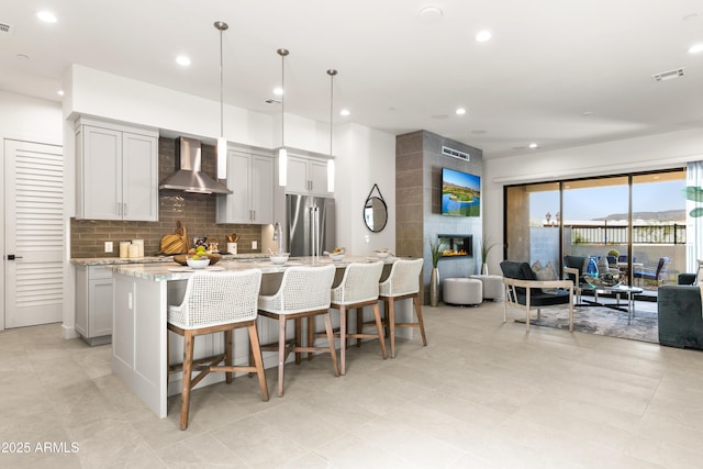 kitchen with pendant lighting, stainless steel fridge, wall chimney range hood, light stone countertops, and a center island with sink