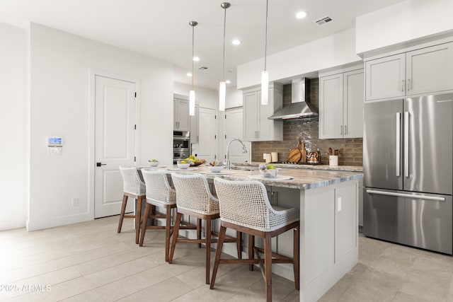 kitchen featuring appliances with stainless steel finishes, a breakfast bar, a kitchen island with sink, light stone counters, and wall chimney range hood