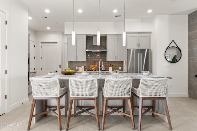 kitchen featuring gray cabinets, stainless steel refrigerator, decorative light fixtures, a kitchen bar, and wall chimney exhaust hood