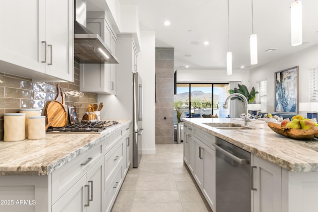 kitchen featuring pendant lighting, wall chimney range hood, appliances with stainless steel finishes, light stone countertops, and a center island with sink