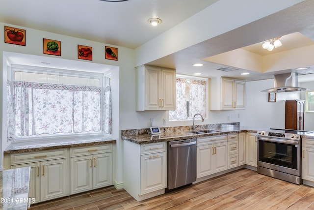 kitchen with appliances with stainless steel finishes, island range hood, light hardwood / wood-style flooring, dark stone counters, and sink