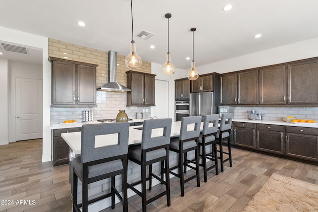 kitchen featuring hanging light fixtures, backsplash, appliances with stainless steel finishes, wall chimney range hood, and a kitchen bar