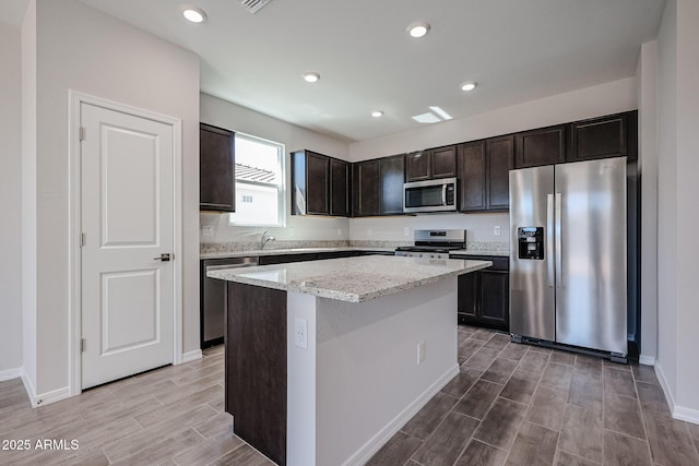 kitchen with appliances with stainless steel finishes, wood tiled floor, dark brown cabinets, light stone countertops, and a kitchen island