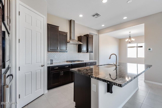 kitchen with sink, a kitchen island with sink, stainless steel gas cooktop, dark stone counters, and wall chimney exhaust hood