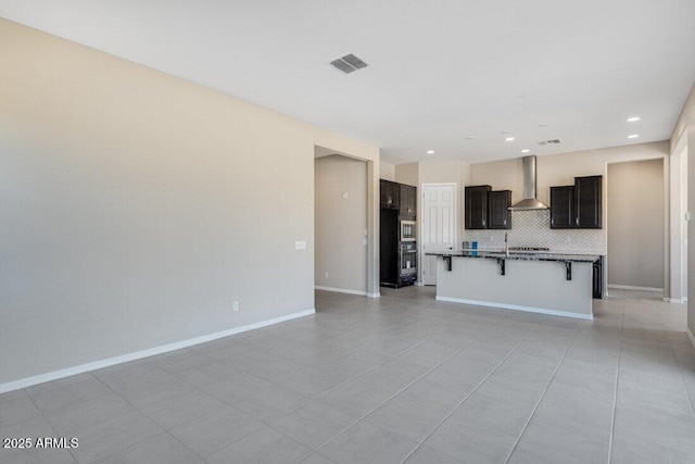 kitchen with wall chimney exhaust hood, a kitchen bar, light tile patterned floors, a kitchen island with sink, and backsplash