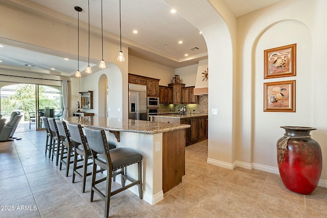 kitchen with arched walkways, a breakfast bar area, light stone countertops, stainless steel built in fridge, and backsplash