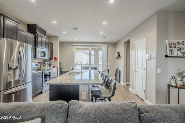 kitchen featuring appliances with stainless steel finishes, a kitchen island with sink, sink, light stone counters, and a breakfast bar area