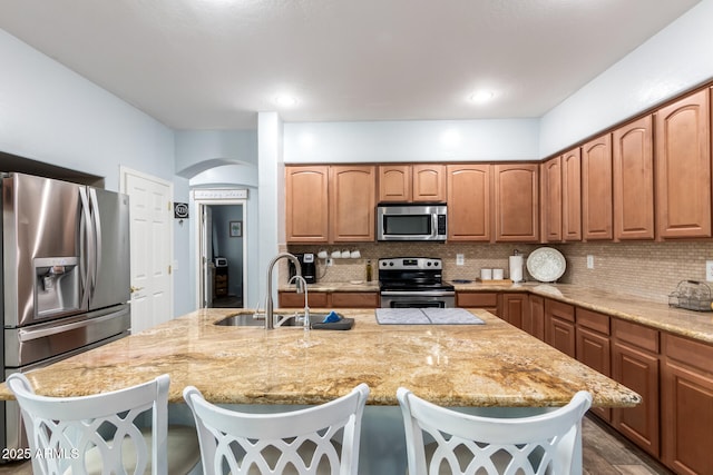 kitchen with stainless steel appliances, decorative backsplash, a sink, an island with sink, and light stone countertops