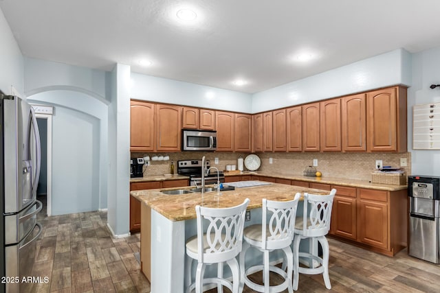 kitchen featuring arched walkways, a kitchen island with sink, a sink, appliances with stainless steel finishes, and dark wood finished floors