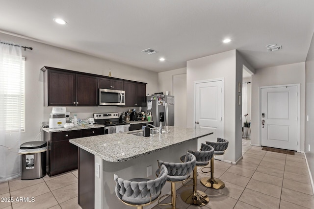 kitchen with light tile patterned floors, appliances with stainless steel finishes, dark brown cabinetry, light stone countertops, and an island with sink