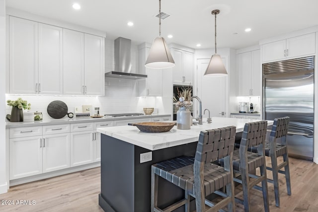 kitchen featuring a kitchen island with sink, light hardwood / wood-style flooring, wall chimney exhaust hood, appliances with stainless steel finishes, and decorative light fixtures