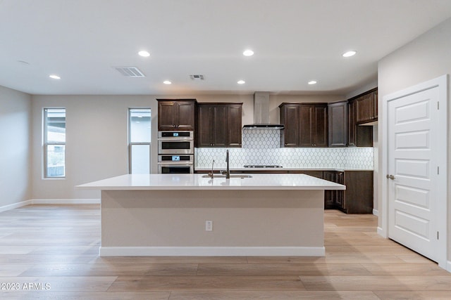 kitchen with appliances with stainless steel finishes, an island with sink, sink, and wall chimney range hood