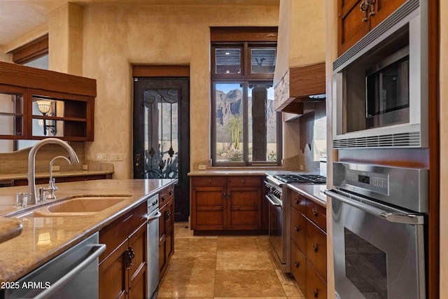 kitchen with open shelves, stainless steel appliances, brown cabinetry, a sink, and light stone countertops