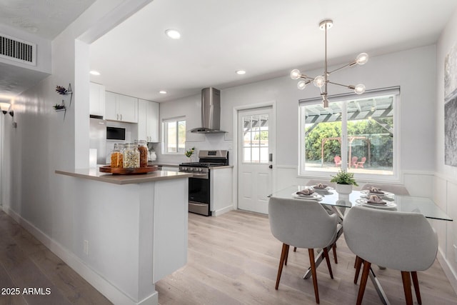 kitchen featuring visible vents, wall chimney range hood, gas range, light wood-style flooring, and a peninsula