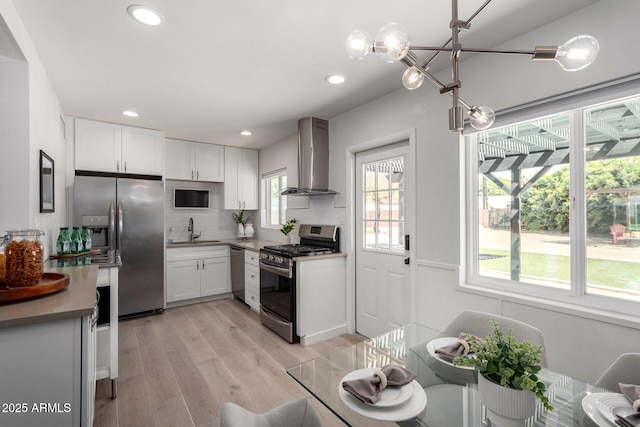 kitchen with wall chimney range hood, light wood-type flooring, white cabinets, stainless steel appliances, and a sink