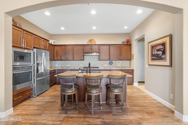 kitchen featuring a breakfast bar area, light stone counters, wood-type flooring, stainless steel appliances, and a kitchen island with sink