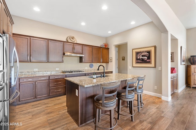 kitchen featuring sink, light wood-type flooring, appliances with stainless steel finishes, an island with sink, and light stone countertops