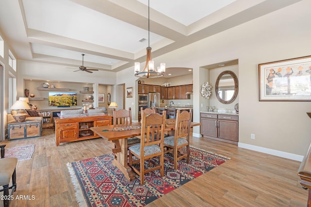 dining space with ceiling fan with notable chandelier, light wood-type flooring, and a tray ceiling