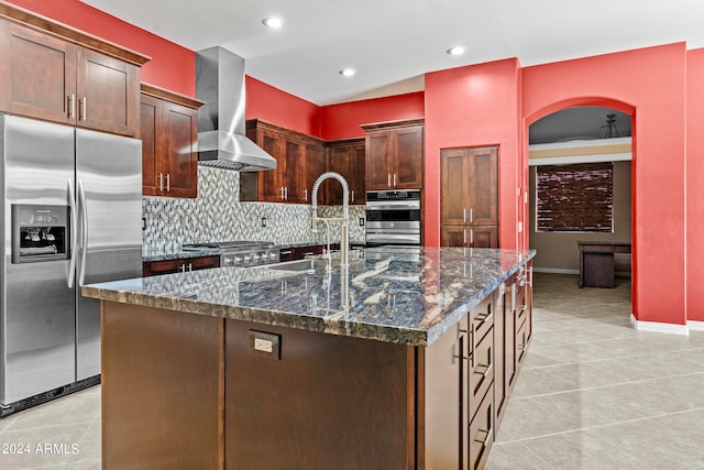 kitchen with appliances with stainless steel finishes, wall chimney range hood, dark stone countertops, an island with sink, and sink