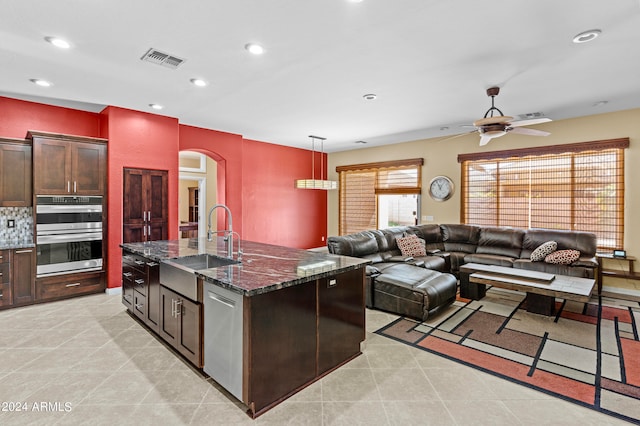 kitchen featuring appliances with stainless steel finishes, a kitchen island with sink, decorative light fixtures, dark stone countertops, and ceiling fan