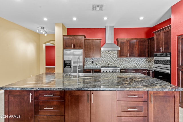 kitchen featuring wall chimney exhaust hood, a kitchen island, stainless steel appliances, dark stone counters, and decorative backsplash