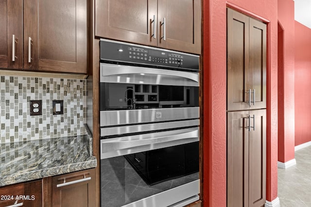 kitchen with light tile patterned floors, double oven, backsplash, dark stone counters, and dark brown cabinetry