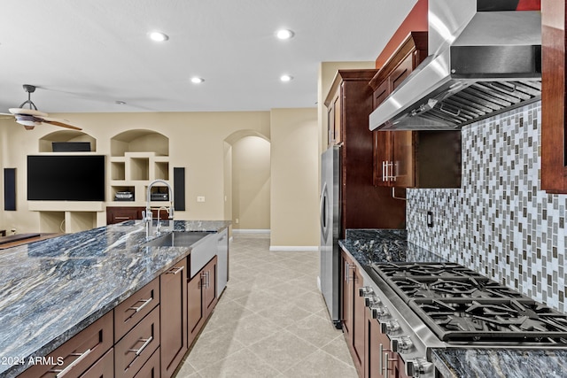 kitchen with baseboards, dark stone counters, appliances with stainless steel finishes, a sink, and wall chimney range hood