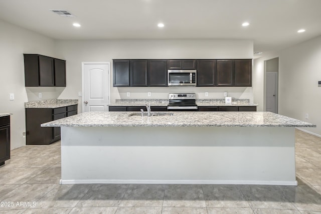 kitchen with light stone countertops, dark brown cabinetry, appliances with stainless steel finishes, sink, and a center island with sink