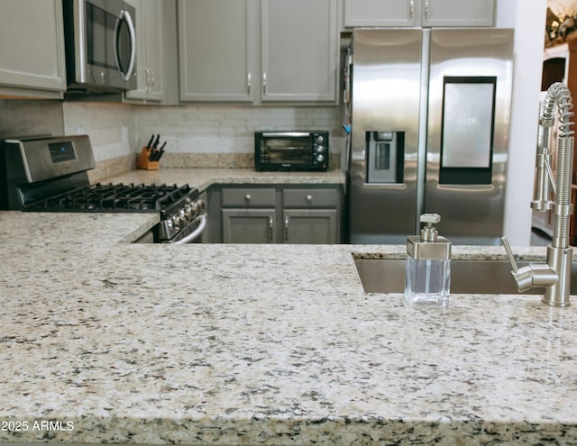 kitchen with backsplash, gray cabinetry, light stone countertops, a toaster, and stainless steel appliances