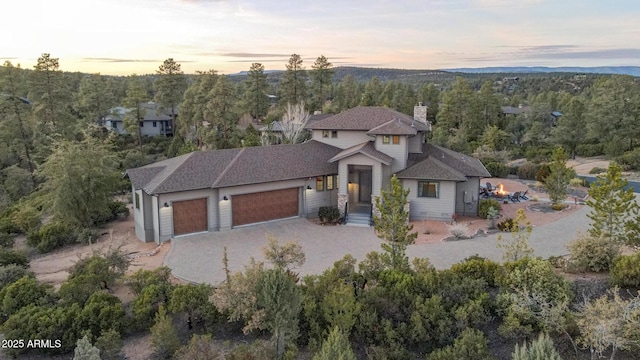 view of front of home with decorative driveway, a chimney, a shingled roof, an attached garage, and a view of trees