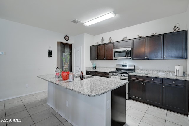 kitchen with sink, a kitchen island with sink, stainless steel appliances, light stone countertops, and dark brown cabinets