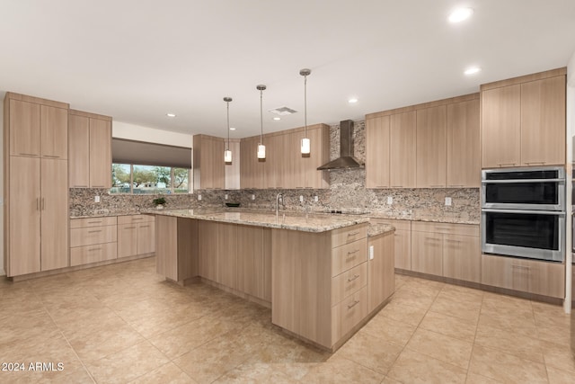 kitchen with stainless steel double oven, wall chimney range hood, an island with sink, and light brown cabinetry