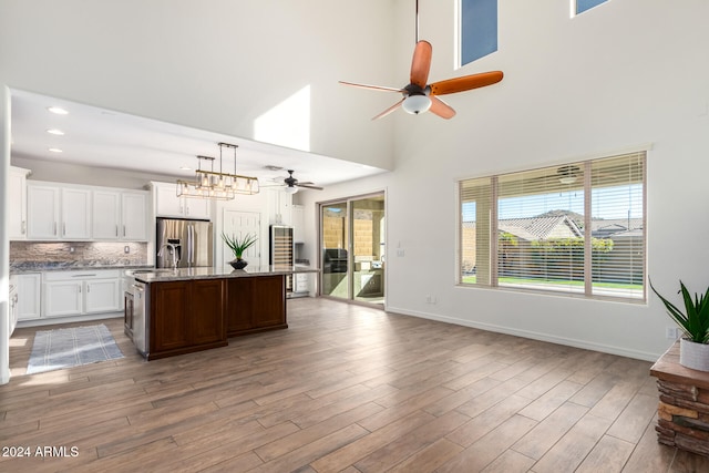 kitchen with hanging light fixtures, a towering ceiling, a kitchen island with sink, stainless steel fridge with ice dispenser, and light wood-type flooring